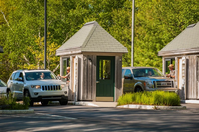 Two small, booth-like buildings are positioned across a roadway. Park rangers are seated at open win